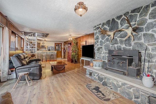 living room with wood walls, a wood stove, ceiling fan, a textured ceiling, and light hardwood / wood-style floors