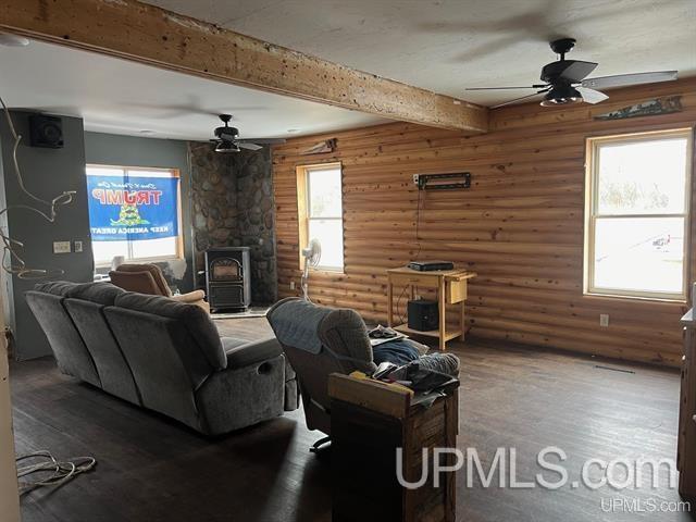 living room featuring ceiling fan, beam ceiling, a wood stove, and log walls