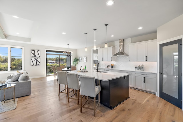 kitchen with white cabinetry, wall chimney range hood, pendant lighting, a kitchen island with sink, and light wood-type flooring