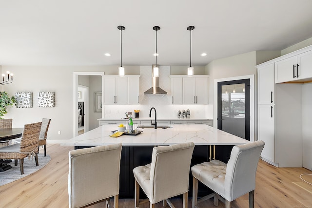 kitchen featuring a kitchen island with sink, sink, hanging light fixtures, wall chimney exhaust hood, and white cabinetry