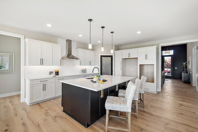 kitchen with a kitchen island with sink, wall chimney range hood, light hardwood / wood-style flooring, white cabinets, and hanging light fixtures