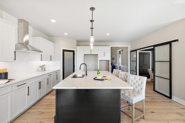 kitchen featuring sink, wall chimney exhaust hood, decorative light fixtures, a center island with sink, and white cabinets