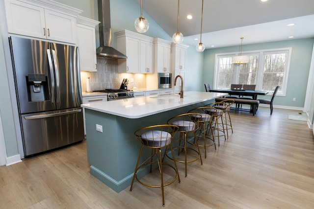 kitchen featuring wall chimney range hood, appliances with stainless steel finishes, white cabinetry, an island with sink, and decorative light fixtures