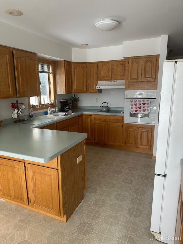 kitchen with brown cabinetry, a sink, a peninsula, white appliances, and under cabinet range hood