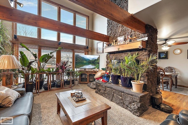 living area featuring ceiling fan, beamed ceiling, a stone fireplace, a high ceiling, and wood finished floors