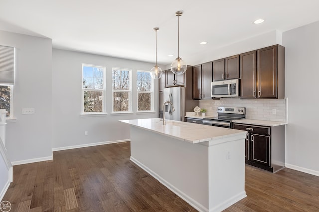 kitchen with appliances with stainless steel finishes, dark hardwood / wood-style flooring, hanging light fixtures, an island with sink, and decorative backsplash