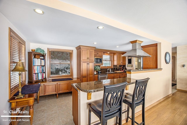 kitchen featuring brown cabinets, a breakfast bar area, stainless steel appliances, dark stone countertops, and island range hood