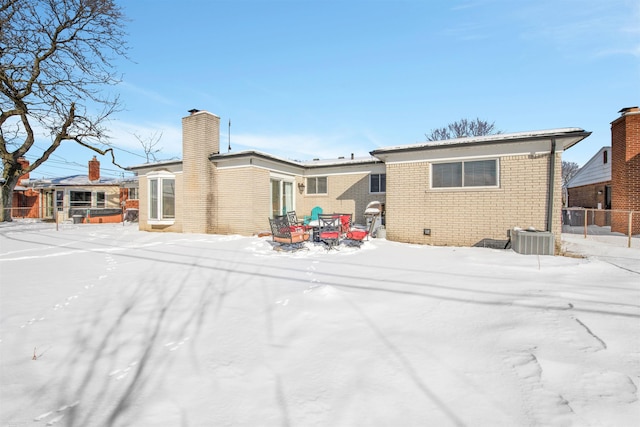 snow covered property featuring brick siding, a chimney, central AC unit, and fence