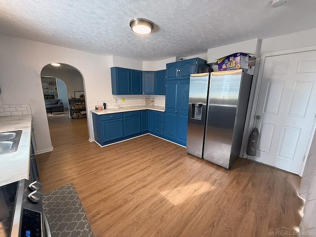 kitchen with arched walkways, blue cabinets, light wood-style floors, light countertops, and stainless steel refrigerator with ice dispenser