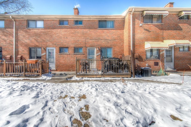 snow covered house with a deck, brick siding, a chimney, and central air condition unit