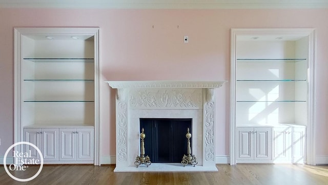 unfurnished living room featuring a fireplace with raised hearth, built in shelves, baseboards, light wood-style floors, and ornamental molding