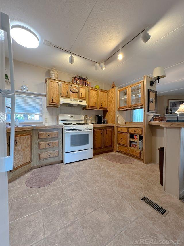 kitchen with white gas range oven, visible vents, brown cabinetry, light countertops, and under cabinet range hood