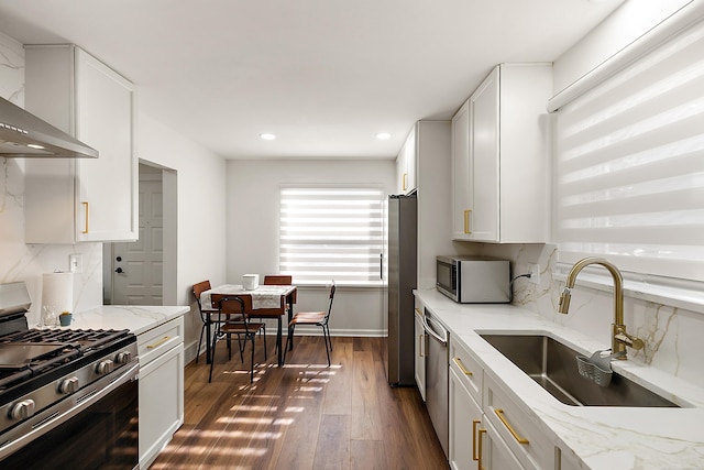 kitchen featuring dark wood finished floors, wall chimney exhaust hood, appliances with stainless steel finishes, light stone countertops, and a sink