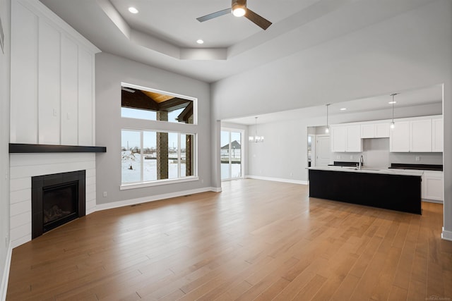 unfurnished living room with a large fireplace, baseboards, light wood-type flooring, a sink, and ceiling fan with notable chandelier