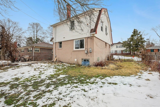 snow covered back of property with central AC unit, brick siding, fence, a gambrel roof, and a chimney