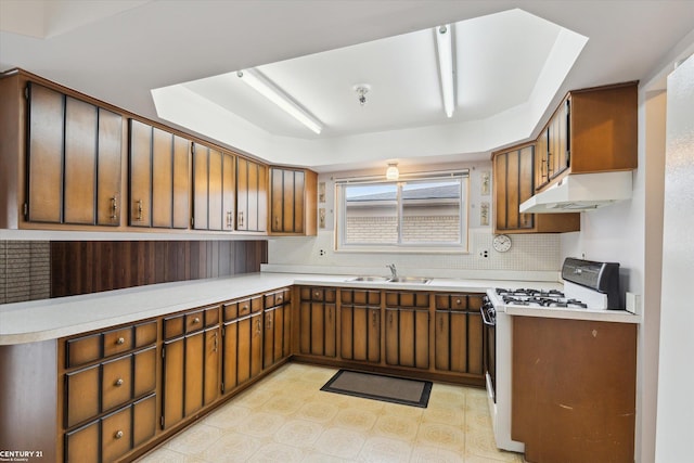 kitchen featuring white gas stove, light floors, backsplash, a sink, and under cabinet range hood