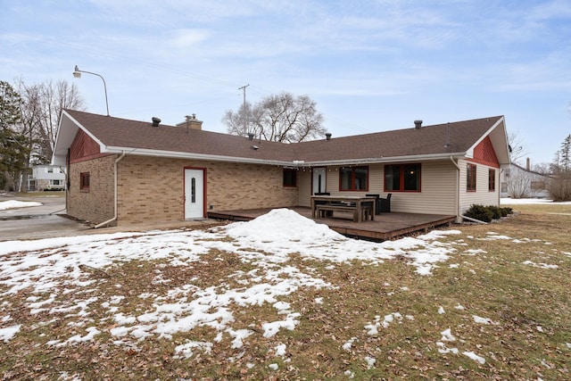 snow covered property with a deck, brick siding, a shingled roof, and a chimney