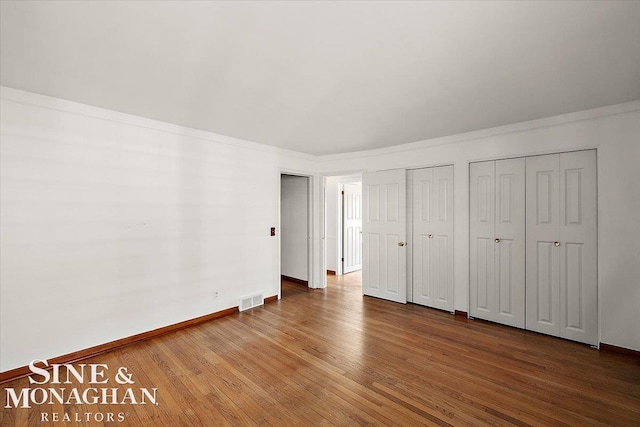 unfurnished bedroom featuring wood finished floors, visible vents, baseboards, multiple closets, and crown molding