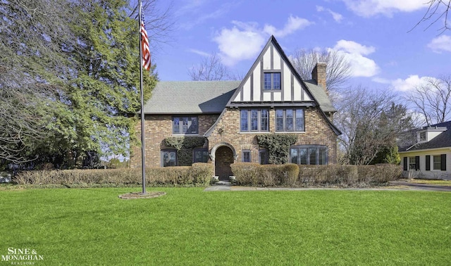 tudor home featuring roof with shingles, a chimney, a front lawn, and brick siding