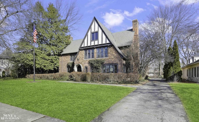 tudor-style house featuring a shingled roof, stucco siding, a chimney, a front yard, and brick siding