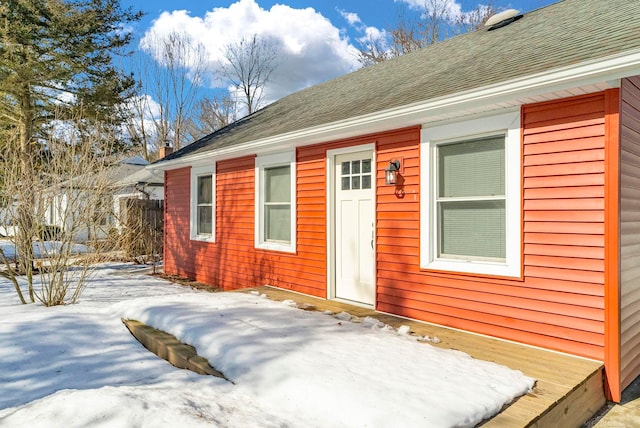 snow covered property entrance featuring roof with shingles and fence