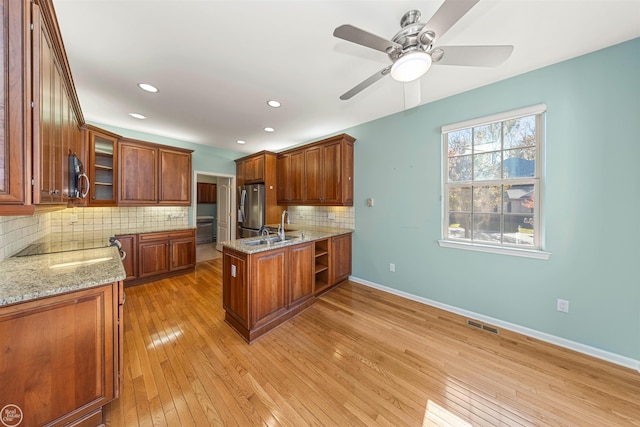 kitchen featuring light wood-type flooring, brown cabinets, appliances with stainless steel finishes, glass insert cabinets, and light stone countertops