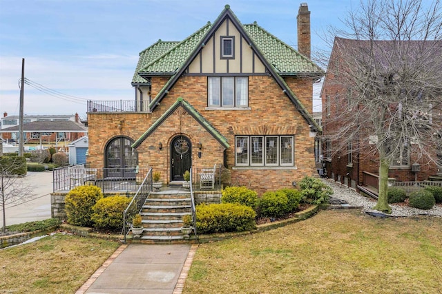 tudor-style house with a balcony, brick siding, a tile roof, a chimney, and a front yard