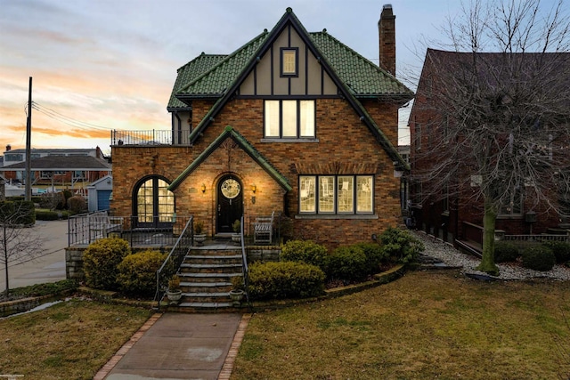 tudor house featuring a balcony, brick siding, a lawn, stucco siding, and a chimney