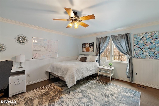 bedroom featuring ceiling fan, a wainscoted wall, wood finished floors, visible vents, and ornamental molding