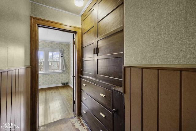 hallway featuring a wainscoted wall, visible vents, ornamental molding, hardwood / wood-style floors, and wallpapered walls