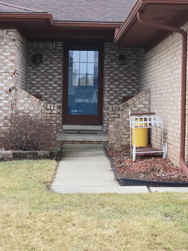 entrance to property with a shingled roof, a lawn, and brick siding