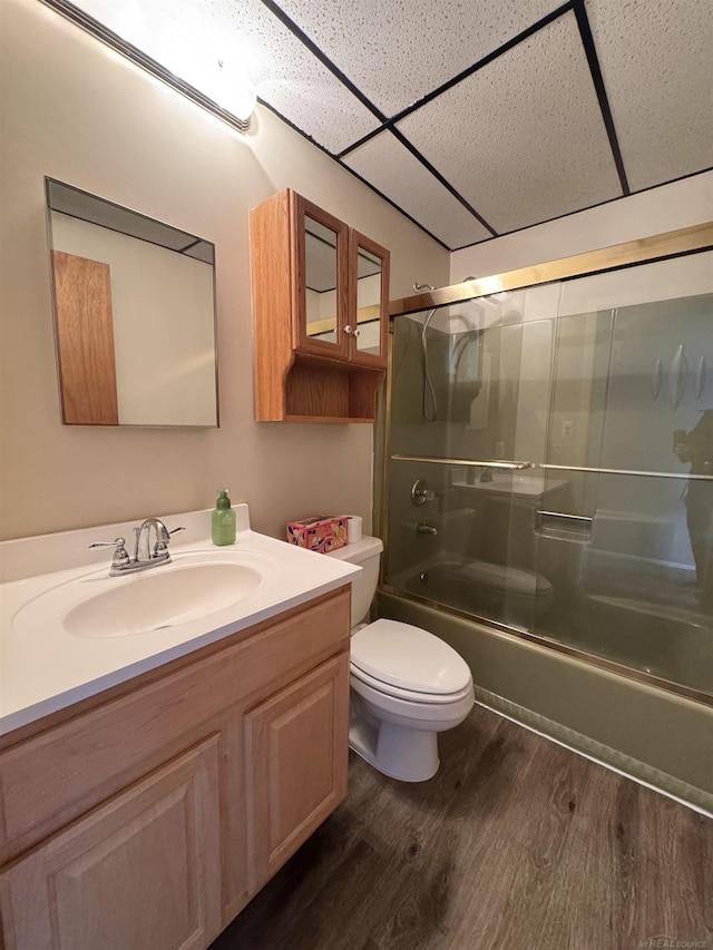 bathroom featuring a drop ceiling, toilet, wood finished floors, combined bath / shower with glass door, and vanity