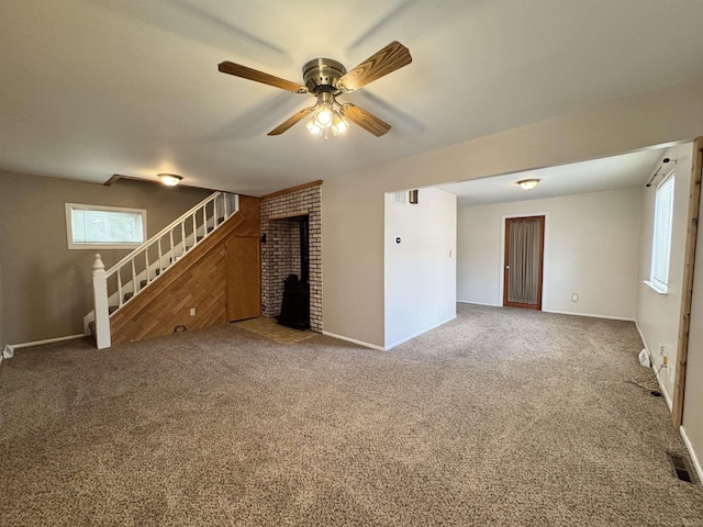 unfurnished living room with visible vents, a ceiling fan, a wood stove, stairs, and carpet floors