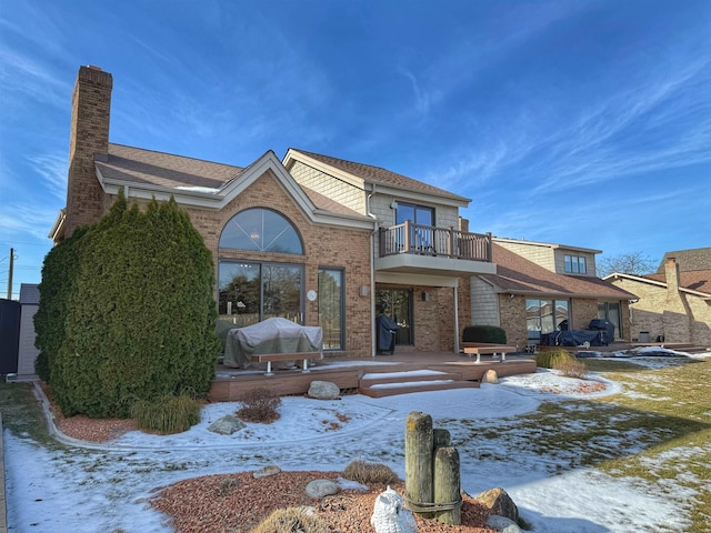 snow covered house featuring a chimney, brick siding, and a balcony