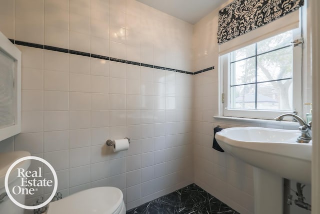 bathroom featuring toilet, marble finish floor, a sink, and tile walls