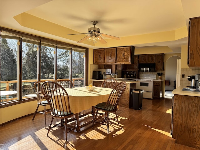 dining area featuring baseboards, arched walkways, a raised ceiling, a ceiling fan, and dark wood-style flooring