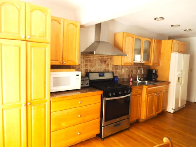 kitchen featuring a sink, backsplash, dark countertops, white appliances, and wall chimney range hood