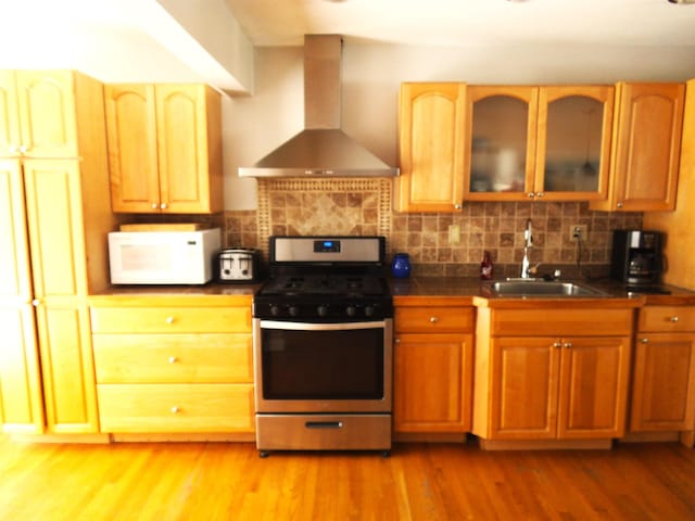 kitchen with wood finished floors, white microwave, a sink, wall chimney exhaust hood, and stainless steel gas stove