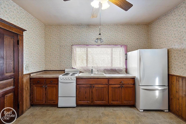 kitchen featuring white appliances, brown cabinetry, a wainscoted wall, wallpapered walls, and a sink