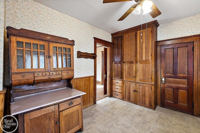 interior space featuring wallpapered walls, a ceiling fan, brown cabinetry, and wainscoting