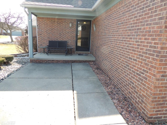 view of exterior entry featuring a patio, brick siding, and roof with shingles