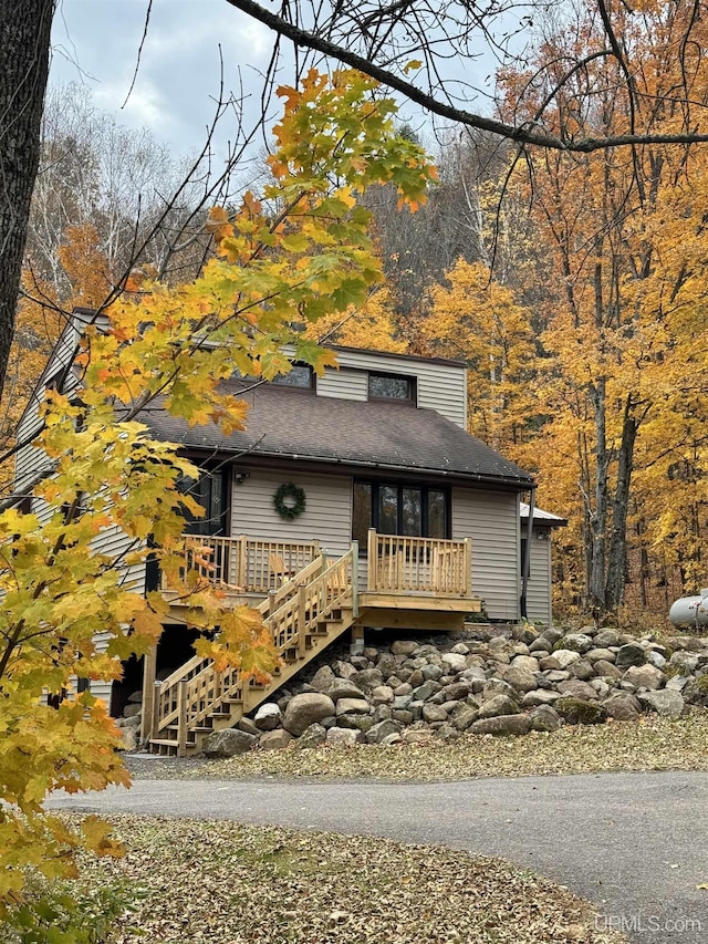 view of front of home with a deck, stairs, a view of trees, and roof with shingles