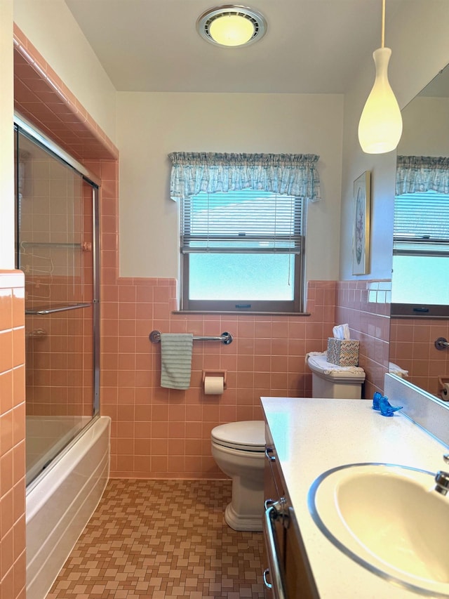 bathroom featuring toilet, shower / bath combination with glass door, tile walls, wainscoting, and vanity