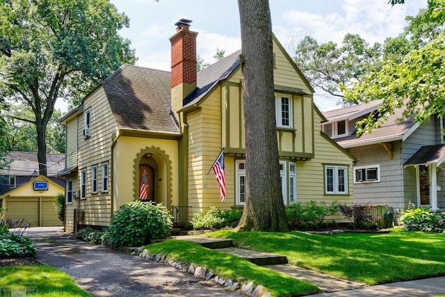 english style home with an outdoor structure, a front yard, a shingled roof, a garage, and a chimney