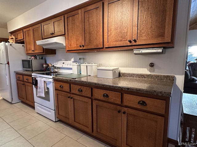 kitchen with dark countertops, under cabinet range hood, brown cabinets, light tile patterned flooring, and white appliances