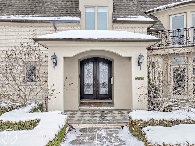 snow covered property entrance with french doors, a balcony, stucco siding, and a shingled roof