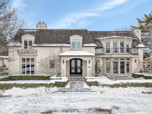 french country style house with brick siding, roof with shingles, french doors, a chimney, and a balcony