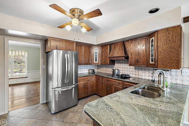 kitchen featuring backsplash, brown cabinetry, freestanding refrigerator, custom exhaust hood, and a sink