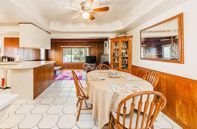 dining room with a ceiling fan, wooden walls, a raised ceiling, and wainscoting