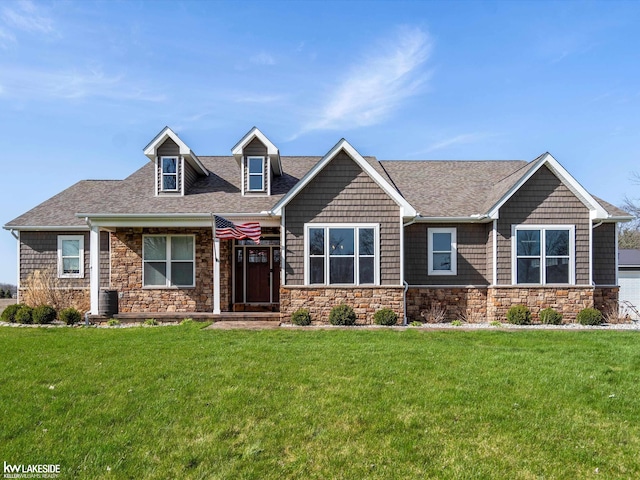 craftsman inspired home featuring cooling unit, a shingled roof, a front yard, and stone siding